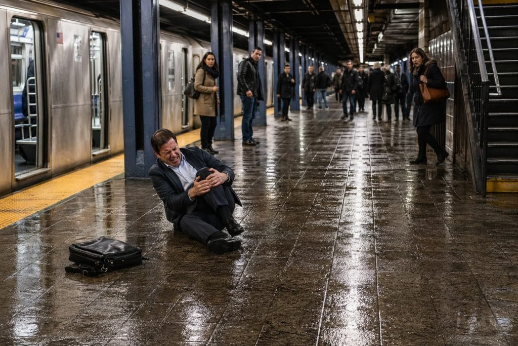 Commuter slipping on a wet NYC subway platform, falling backward while clutching their knee as nearby passengers react in a busy transit setting.